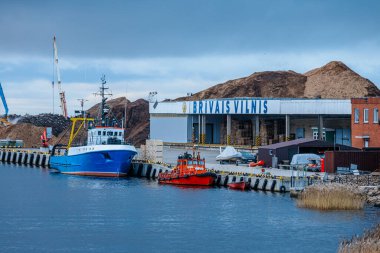 Bugoe adında büyük kırmızı bir kargo gemisi bulutlu bir gökyüzünün altında görünür cankurtaran botları ve odun yığınları olan bir limana yanaştı. Denizcilik, ticaret ve endüstriyel temalar için ideal. 11.19.2024. Selâm, Letonya