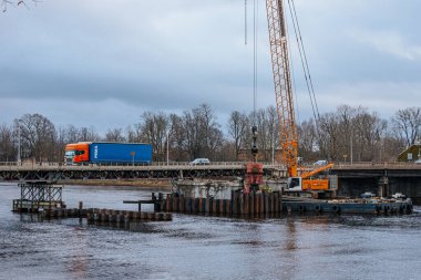 Bugoe adında büyük kırmızı bir kargo gemisi bulutlu bir gökyüzünün altında görünür cankurtaran botları ve odun yığınları olan bir limana yanaştı. Denizcilik, ticaret ve endüstriyel temalar için ideal. 11.19.2024. Selâm, Letonya