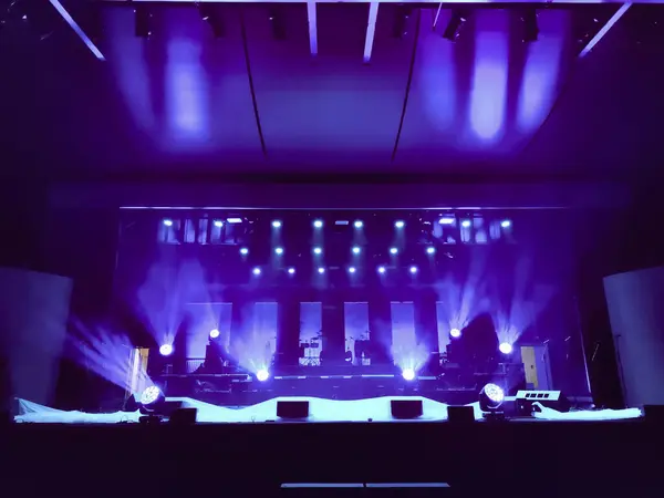 Sahne ışıkları, müzik ekipmanları ve dramatik bir atmosfer içeren canlı mavi ve mor ışıklarla aydınlatılmış bir konser sahnesi. Etkinlik terfileri için mükemmel.