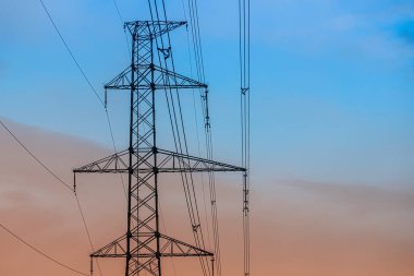 Yüksek voltajlı bir elektrik direği bulutlu mavi bir gökyüzüne karşı duruyor ve çoklu elektrik hatlarını destekliyor. Çelik kafes yapısı endüstriyel enerji altyapısını vurguluyor.
