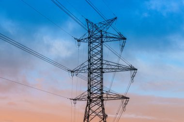 Yüksek voltajlı bir elektrik direği bulutlu mavi bir gökyüzüne karşı duruyor ve çoklu elektrik hatlarını destekliyor. Çelik kafes yapısı endüstriyel enerji altyapısını vurguluyor.