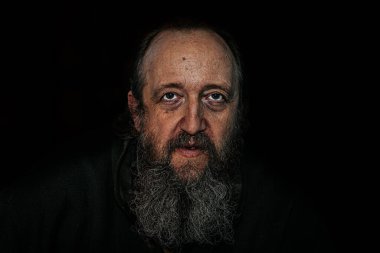 Derin kırışıklıkları, uzun sakalı ve delici gözleri olan yaşlı bir adamın dramatik yakın plan portresi. Karanlık arka plan, yoğun ve duygusal ifadeyi güçlendirir..