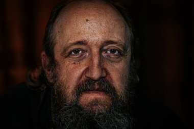 Derin kırışıklıkları, uzun sakalı ve delici gözleri olan yaşlı bir adamın dramatik yakın plan portresi. Karanlık arka plan, yoğun ve duygusal ifadeyi güçlendirir..