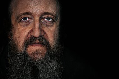 Derin kırışıklıkları, uzun sakalı ve delici gözleri olan yaşlı bir adamın dramatik yakın plan portresi. Karanlık arka plan, yoğun ve duygusal ifadeyi güçlendirir..