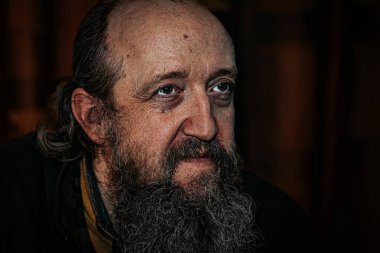 Derin kırışıklıkları, uzun sakalı ve delici gözleri olan yaşlı bir adamın dramatik yakın plan portresi. Karanlık arka plan, yoğun ve duygusal ifadeyi güçlendirir..
