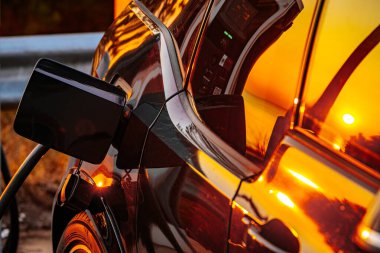Şarj istasyonuna takılı elektrikli bir aracın yakın plan görüntüsü, gün batımının parlak siyah yüzeyine yansıması. Sürdürülebilir ulaşım ve temiz enerjinin sembolü.