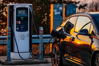 Şarj istasyonuna takılı elektrikli bir aracın yakın plan görüntüsü, gün batımının parlak siyah yüzeyine yansıması. Sürdürülebilir ulaşım ve temiz enerjinin sembolü.