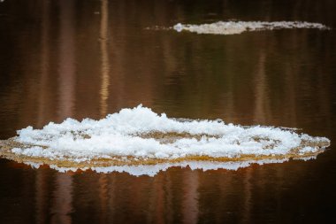 Yüzen bir buz parçası sakin bir nehirde sürükleniyor, karanlık suları yansıtıyor. Katmanlı buz oluşumu arka plandaki sıcak tonlarla çelişiyor. Sakin bir kış sahnesi.