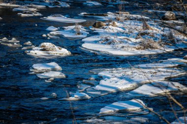 Karla kaplı kıyılar ve buzla kaplı ağaçlarla çevrili yumuşak akan bir nehrin olduğu huzurlu bir kış manzarası. Parlak güneş ışığı, kışın doğanın keskin güzelliğini vurgular..