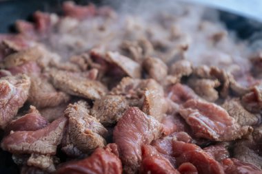 Taze kesilmiş et sıcak bir tavada cızırdıyor ve et pişerken buhar yükseliyor. Çiğ ve kahverengi parçaların karışımı yemek pişirme sürecini ve zengin tatların salınımını vurguluyor..