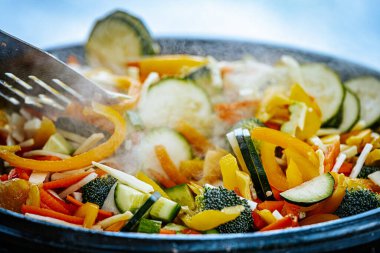 A steaming vegetable stir-fry with zucchini, bell peppers, carrots, and broccoli is being cooked in a pan. A spatula is stirring the fresh, colorful ingredients. clipart