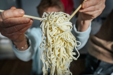Bir insan yemek çubuklarıyla pişmiş eriştenin bir kısmını kaldırır, dokusunu ve esnekliğini gösterir. Sahne, geleneksel bir Asya yemek deneyimini vurguluyor.