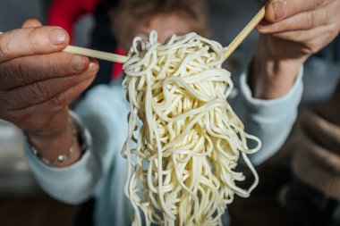 Bir insan yemek çubuklarıyla pişmiş eriştenin bir kısmını kaldırır, dokusunu ve esnekliğini gösterir. Sahne, geleneksel bir Asya yemek deneyimini vurguluyor.