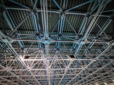 Endüstriyel metal bir tavanın detaylı bir görüntüsü ve karmaşık bir şebeke yapısı. Mavi ışıklandırma, modern bir tesiste güçlü bir çerçeve, mühendislik ve mimari tasarımı ön plana çıkarıyor.