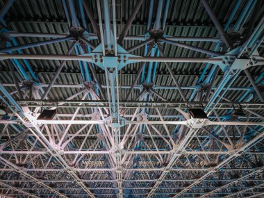 Endüstriyel metal bir tavanın detaylı bir görüntüsü ve karmaşık bir şebeke yapısı. Mavi ışıklandırma, modern bir tesiste güçlü bir çerçeve, mühendislik ve mimari tasarımı ön plana çıkarıyor.