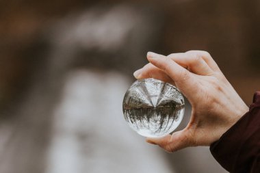 Bir el, bir orman yolunun ters görüntüsünü yansıtan şeffaf bir cam küreyi tutar. Bulanık arkaplan kompozisyonun sanatsal ve soyut doğasını geliştirir.