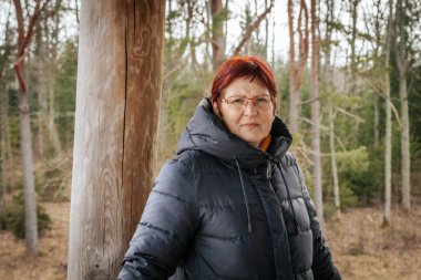 Siyah kışlık ceket giyen, kızıl saçlı ve gözlüklü yaşlı bir kadının portresi. Ciddi ifadesi bulanık orman geçmişiyle çelişiyor. Doğal ve düşünceli bir atmosfer yaratıyor..