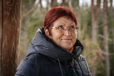 Siyah kışlık ceket giyen, kızıl saçlı ve gözlüklü yaşlı bir kadının portresi. Ciddi ifadesi bulanık orman geçmişiyle çelişiyor. Doğal ve düşünceli bir atmosfer yaratıyor..