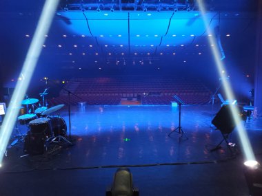 Rows of empty chairs face a stage illuminated by dramatic blue and white lighting. The setup suggests anticipation for an upcoming event or performance in a concert hall.