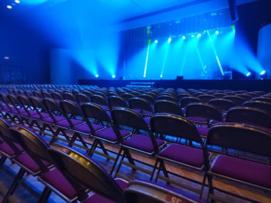 Rows of empty chairs face a stage illuminated by dramatic blue and white lighting. The setup suggests anticipation for an upcoming event or performance in a concert hall.