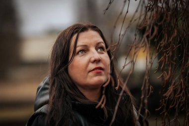 A woman in a black hooded jacket leans against a tree, gazing into the distance. The blurred forest background and overcast lighting create a moody and contemplative atmosphere.