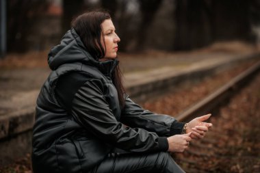 A woman in a black leather outfit and high-heeled boots stands on railway tracks, extending her hand while gazing back. The moody setting and overcast sky create a sense of mystery.