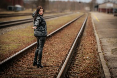 A woman in a black leather outfit and high-heeled boots stands on railway tracks, extending her hand while gazing back. The moody setting and overcast sky create a sense of mystery.