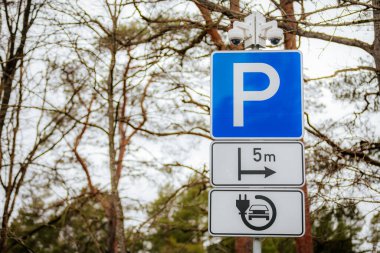 Üzerinde beyaz P işareti olan mavi bir park yeri işareti, belirlenmiş bir park yerini gösteriyor. Arkaplan bulanık, ağaç dallarını ve bulutlu gökyüzünü gösteriyor.