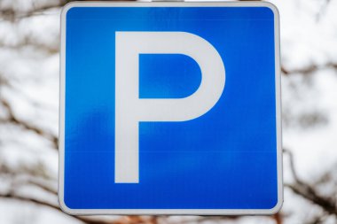 Üzerinde beyaz P işareti olan mavi bir park yeri işareti, belirlenmiş bir park yerini gösteriyor. Arkaplan bulanık, ağaç dallarını ve bulutlu gökyüzünü gösteriyor.