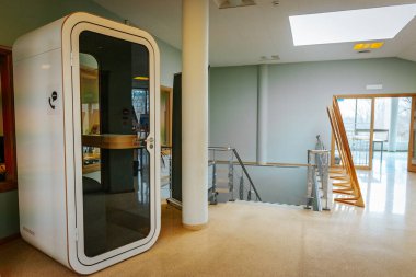 Camdan kapısı olan modern bir ofis telefonu kulübesi, kapalı bir alanda duruyor. Bölgede minimalist dekorasyon, büyük pencereler ve ahşap elementler yer alıyor. 03.09.2025 Valmiera, Letonya