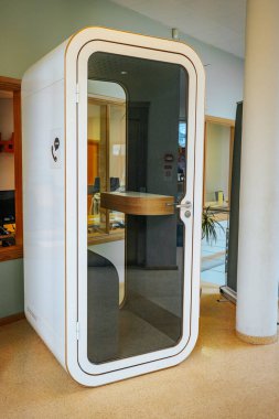 Camdan kapısı olan modern bir ofis telefonu kulübesi, kapalı bir alanda duruyor. Bölgede minimalist dekorasyon, büyük pencereler ve ahşap elementler yer alıyor. 03.09.2025 Valmiera, Letonya