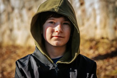 Siyah kapüşonlu, yeşil kapüşonlu bir çocuk ormandaki bir kaya çıkıntısında oturur ve sakince uzaklığa bakar. Ağaçların arasından süzülen sıcak ışık huzurlu ve rahat bir atmosfer yaratıyor..