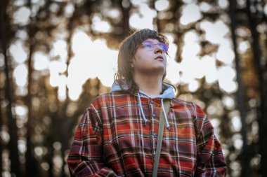 Gözlük takan, ekose ceket giyen ve kapüşonlu giyen genç bir adam derin düşüncelere dalmış bir ifadeyle yukarı bakıyor. Sıcak güneş ışığı ağaçların arasından süzülerek rüya gibi bir atmosfer yaratır..