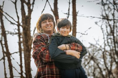 Ekose ceketli gülümseyen bir genç siyah kapüşonlu bir çocuğu kucaklıyor, arka planda çıplak ağaçlar ve yumuşak güneş ışığı olan açık hava ortamında neşeli bir anı paylaşıyor..