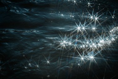 Güneş ışığı dalgalanan sudan yansıyarak büyüleyici bir yıldız patlaması etkisi yaratır. Parlak, kesişen ışık ışınları su yüzeyinde rüya gibi ve soyut bir desen oluşturur..