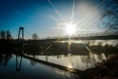 Asma köprü, berrak mavi gökyüzünün altındaki sakin bir nehri kaplar ve güneş bir yıldız patlaması etkisi yaratır. Su, köprüyü ve etrafındaki ağaçları yansıtıyor ve sakin manzarayı güçlendiriyor..