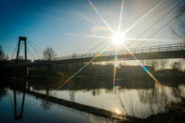 Asma köprü, berrak mavi gökyüzünün altındaki sakin bir nehri kaplar ve güneş bir yıldız patlaması etkisi yaratır. Su, köprüyü ve etrafındaki ağaçları yansıtıyor ve sakin manzarayı güçlendiriyor..