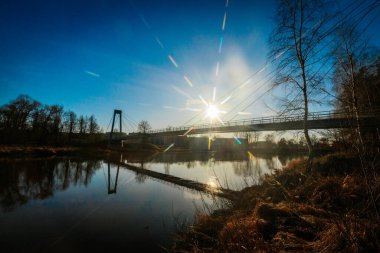 Asma köprü, berrak mavi gökyüzünün altındaki sakin bir nehri kaplar ve güneş bir yıldız patlaması etkisi yaratır. Su, köprüyü ve etrafındaki ağaçları yansıtıyor ve sakin manzarayı güçlendiriyor..