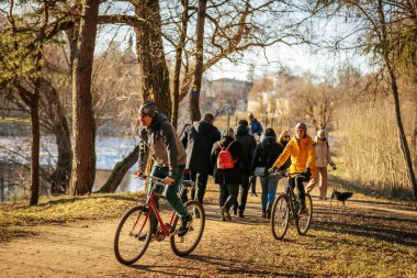 İnsanlar parkta güneşli bir günün tadını çıkarıyorlar. Ağaçlarla kaplı manzaralı bir patika boyunca bisiklet sürüp yürüyorlar. Sıcak güneş ışığı açık hava atmosferini canlandırır. 03.09.2025 Valmiera, Letonya