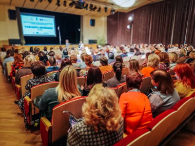 Kalabalık bir seyirci, iyi aydınlatılmış bir konferans salonunda sunum dinliyor. Olay, bilgileri gösteren projektör ekranlı bir sahne içeriyor. 03.12.2025 Valmiera, Letonya