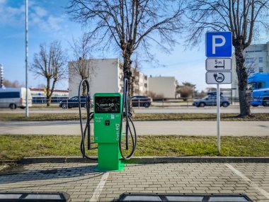 Birden fazla kablosu olan yeşil bir elektrikli araç şarj istasyonu belirlenmiş park alanında yer almaktadır. Arka planda bir park yeri ve şehir binaları görünüyor. 03.12.2025 Valmiera, Letonya