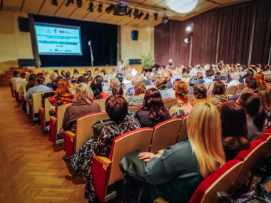 Kalabalık bir seyirci, iyi aydınlatılmış bir konferans salonunda sunum dinliyor. Olay, bilgileri gösteren projektör ekranlı bir sahne içeriyor. 03.12.2025 Valmiera, Letonya