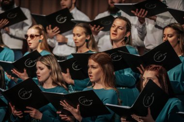 Bir orkestra şefinin yönetiminde sahnedeki canlı performansları senkronize ve tutkulu bir şekilde müzik kağıtları tutan bir koro şefinin yönetiminde sahneye çıkıyor. 03.15.2025 Valmiera, Letonya