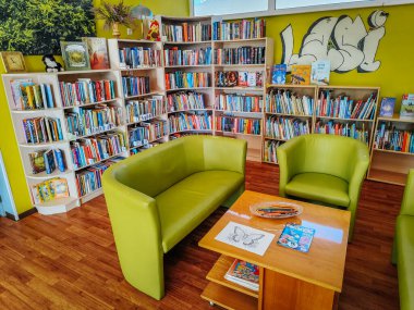Renkli kitaplıklarla dolu parlak ve davetkar bir kütüphane odası, yeşil bir kanepe ve koltuklar, ve çizim malzemeleri olan ahşap bir masa. 03.15.2025 Staicele, Letonya