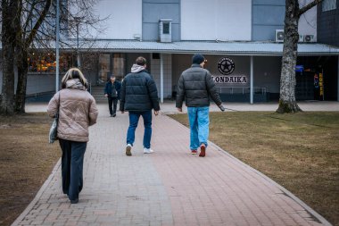 Mavi ceketli ve şapkalı yaşlı bir adam bir cüzdan tutarken asfalt bir yolda yürüyor. Arka planda, bir anne ve çocuk da dahil olmak üzere insanlar yürüyorlar. 03.20.2025 Valmiera, Letonya