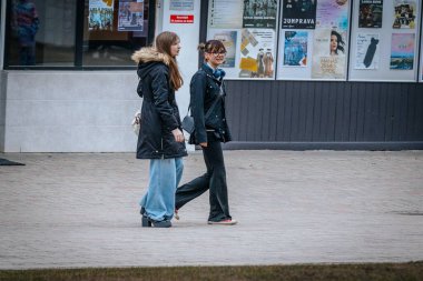 İki genç kadın şehir kaldırımında birlikte yürüyor, günlük ve şık kıyafetler giyiyorlar. Biri kürklü siyah bir palto giyerken, diğerinin kulaklığı 0.03.20.2025 civarında. Valmiera, Letonya