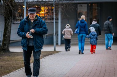 Mavi ceketli ve şapkalı yaşlı bir adam bir cüzdan tutarken asfalt bir yolda yürüyor. Arka planda, bir anne ve çocuk da dahil olmak üzere insanlar yürüyorlar. 03.20.2025 Valmiera, Letonya