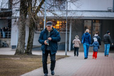 Mavi ceketli ve şapkalı yaşlı bir adam bir cüzdan tutarken asfalt bir yolda yürüyor. Arka planda, bir anne ve çocuk da dahil olmak üzere insanlar yürüyorlar. 03.20.2025 Valmiera, Letonya