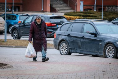 Eskimiş bordo ceketli yaşlı bir adam elinde plastik bir torbayla bir şehir caddesinde yürüyor. Yorgun bir ifadeyle evsiz görünüyor ve park halindeki arabalarla çevrili. 03.20.2025 Zina, Letonya