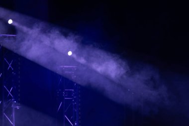 Mor sahne ışıkları sisli sisi delip geçer, karanlık bir tiyatro ortamında metal kirişleri vurgular. Güçlü atmosfer ve aydınlatma efektleri.